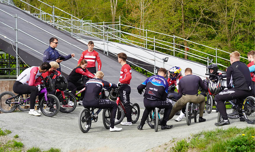 A group of people wearing cycling gear and helmets are gathered on BMX bikes on a dirt track while a person in casual clothing appears to be giving instructions. Description generated by AI