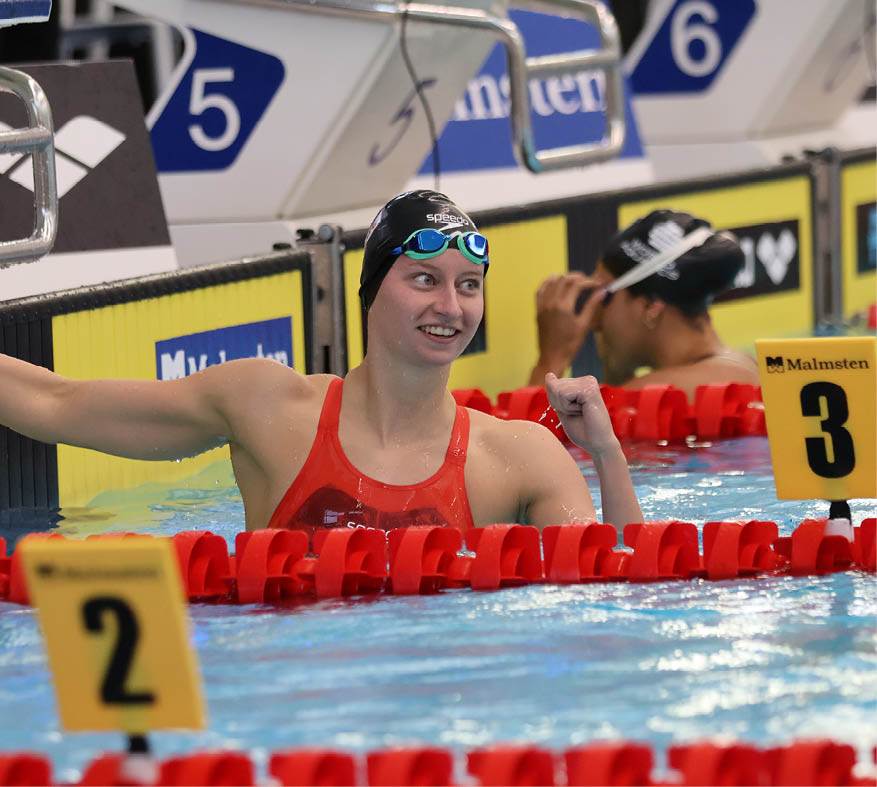 A person wearing a swim cap and goggles is smiling and raising a fist in celebration while resting at the edge of a swimming pool near a lane marker. Description generated by AI